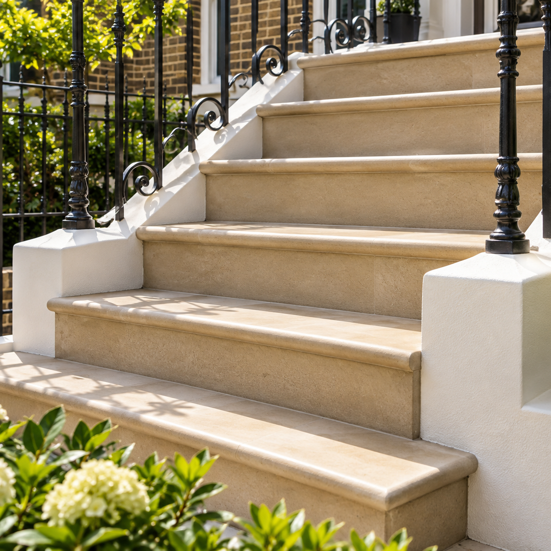 A set of elegant outdoor stone steps with a warm beige finish, flanked by white pedestals and black wrought iron railings, set in a lush garden with green foliage and white flowers.