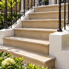 A set of elegant outdoor stone steps with a warm beige finish, flanked by white pedestals and black wrought iron railings, set in a lush garden with green foliage and white flowers.