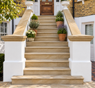 A set of elegant outdoor stone steps leading up a front porch, flanked by white painted posts with warm beige stone railings and potted plants.